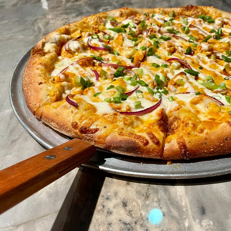 A Lucy Blue Pizza with chicken, red onions, and green onions, on a metal pizza pan.