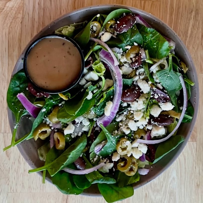 Overhead view of a fresh spinach salad with olives, red onion, feta, and dressing on a wooden table.