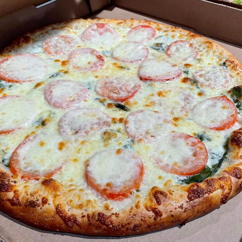 A whole Lucy Blue Pizza with melted cheese, tomato slices, and spinach in a delivery box.
