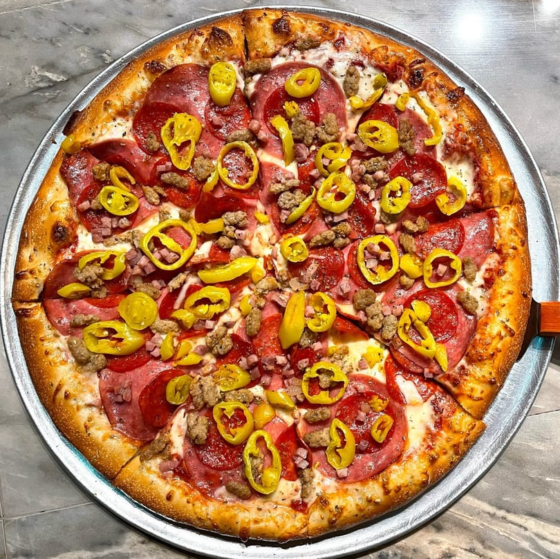 A Lucy Blue Pizza with pepperoni, sausage, ham, and yellow banana peppers on a metal tray.