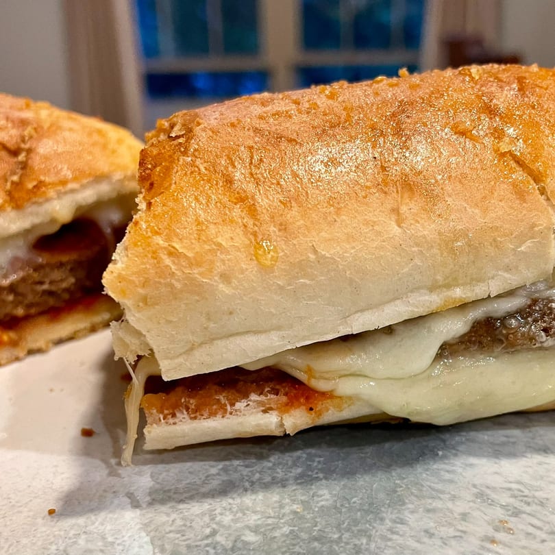 Two Lucy Blue Pizza meatball subs with melted cheese and sauce on a white plate.