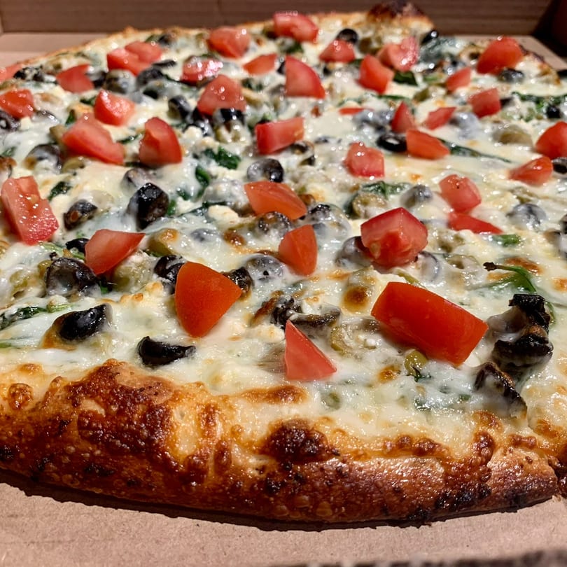 Lucy Blue Pizza with white sauce, black olives, spinach, and diced tomatoes in a box.